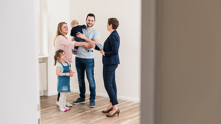 Zu sehen ist eine Familie mit zwei Kindern mit einer Maklerin in einer leeren Wohnung